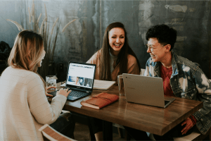Three colleagues laughing and collaborating while working on laptops at a wooden table.