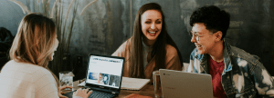 Three colleagues laughing and collaborating while working on laptops at a wooden table.