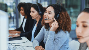 Smiling female call center agent wearing a headset, assisting a client in a vibrant, modern call center with other female colleagues.